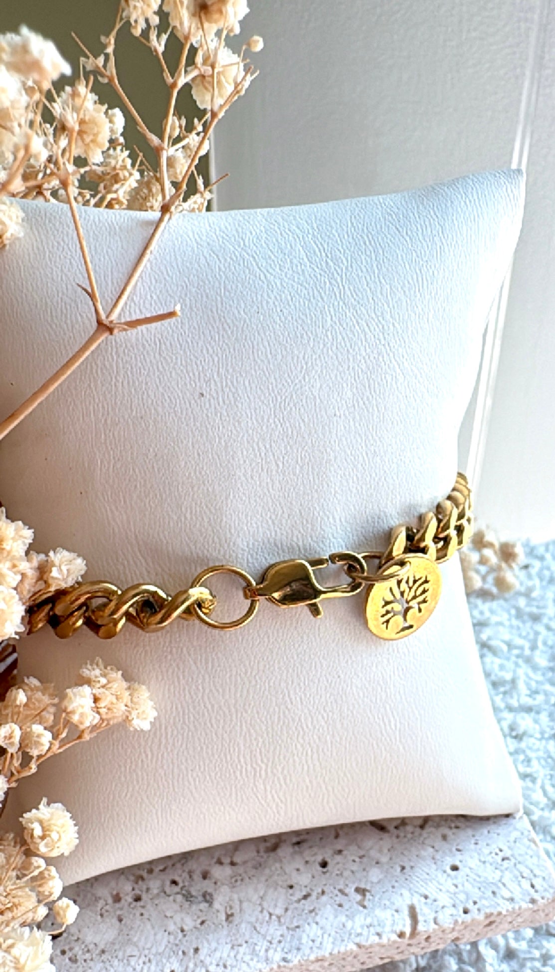 Gold bracelet with an engraved tree  in a circular gold pendant on a white cushion with dried flowers