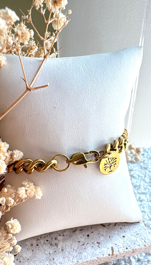 Gold bracelet with an engraved tree  in a circular gold pendant on a white cushion with dried flowers